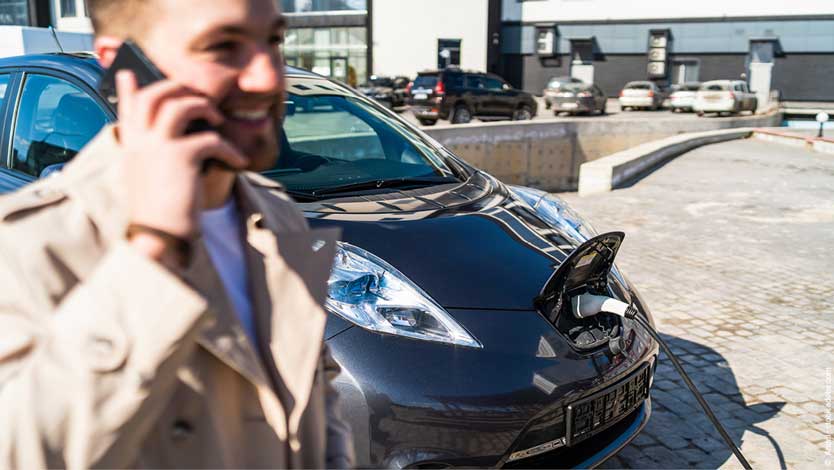 Ein Mann steht an der Tankstelle und telefoniert, während er sein E-Auto auflädt. Diese Autos helfen bei der technologieneutralen Erreichung der Klimaziele.