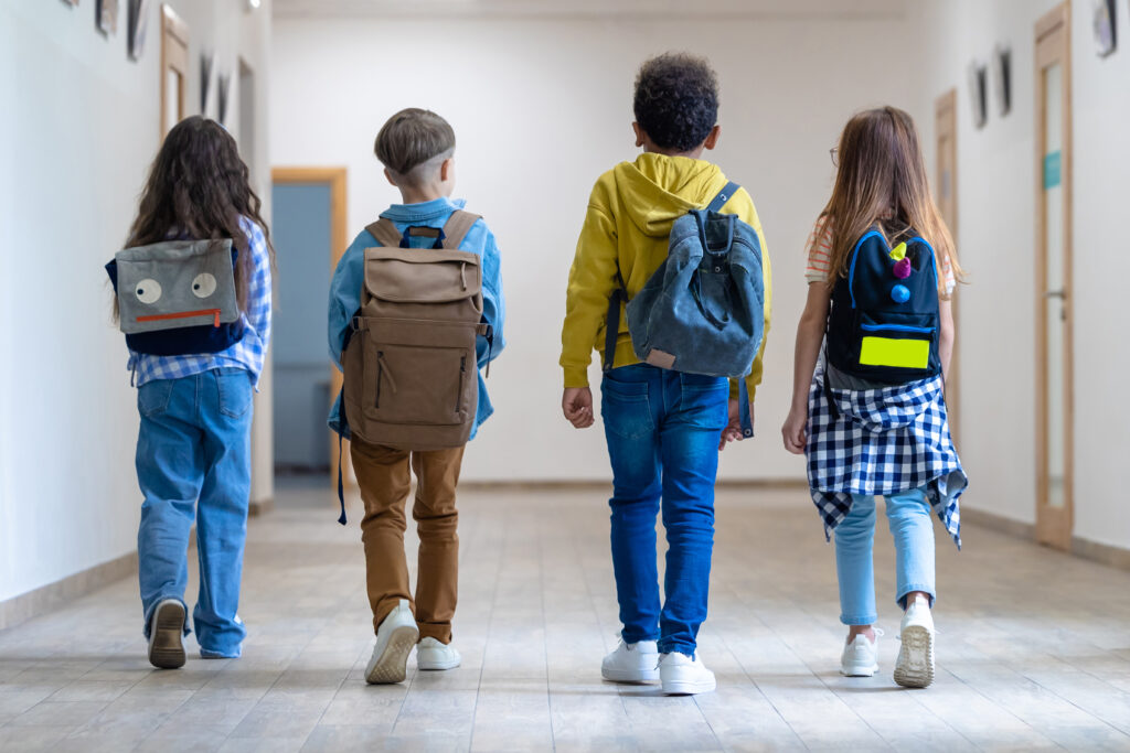Vier Kinder mit Schultaschen gehen von hinten zu sehen nebeneinander durch einen Gang