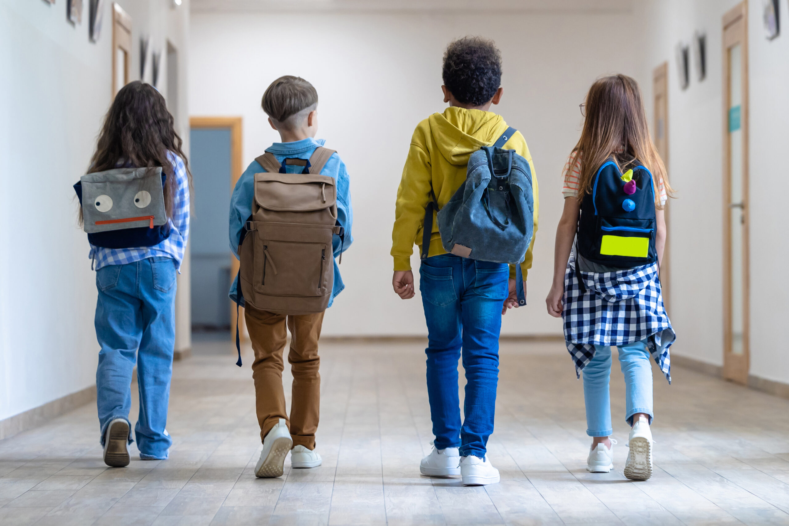 Vier Kinder mit Schultaschen gehen von hinten zu sehen nebeneinander durch einen Gang